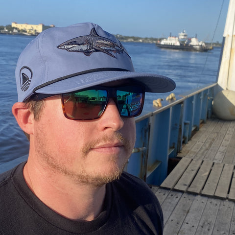 Man standing on boat wearing dusty blue hat with shark graphic on front and Ocearch logo on side