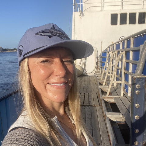 Woman standing on boat wearing dusty blue hat with shark graphic on front and Ocearch logo on side