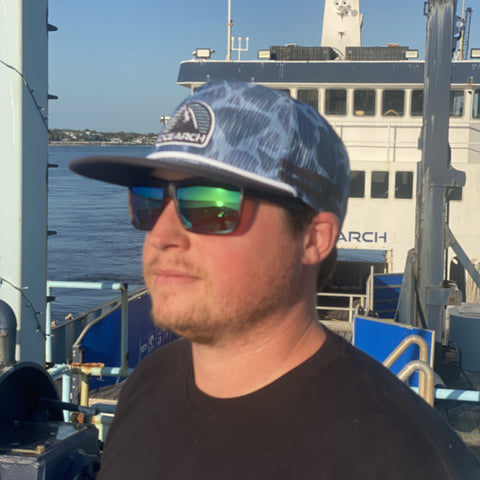Man standing on boat wearing camo blue mesh hat with "OCEARCH" patch on front